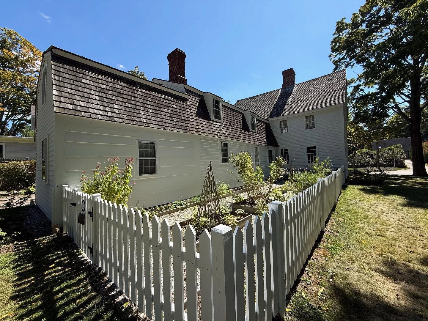 White house with brown shingle roof, chimney, and picket fence garden on a sunny day.