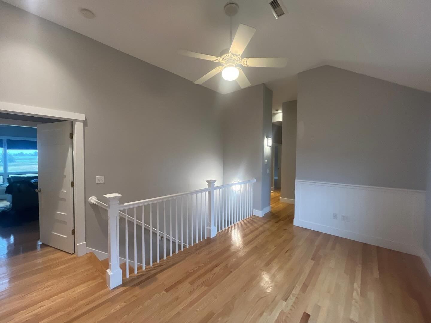 A bright, airy second-story hallway with hardwood floors, a white railing, and a gray wall; a ceiling fan is visible.