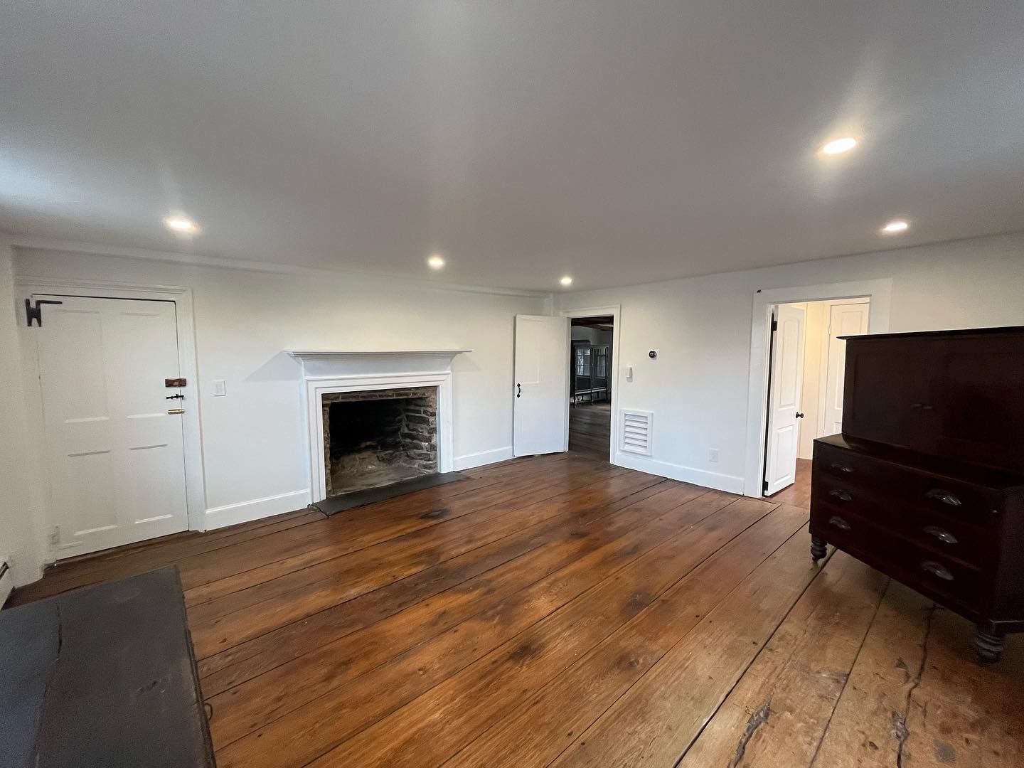 Spacious room with fireplace, hardwood floors, and a wooden dresser. White walls and doors, bright overhead lights.