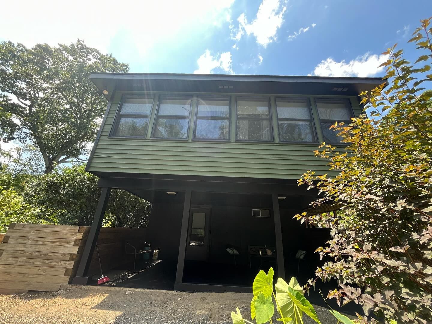 Green, two-story house with windows. Underneath is an open space. Trees surround it on a sunny day.