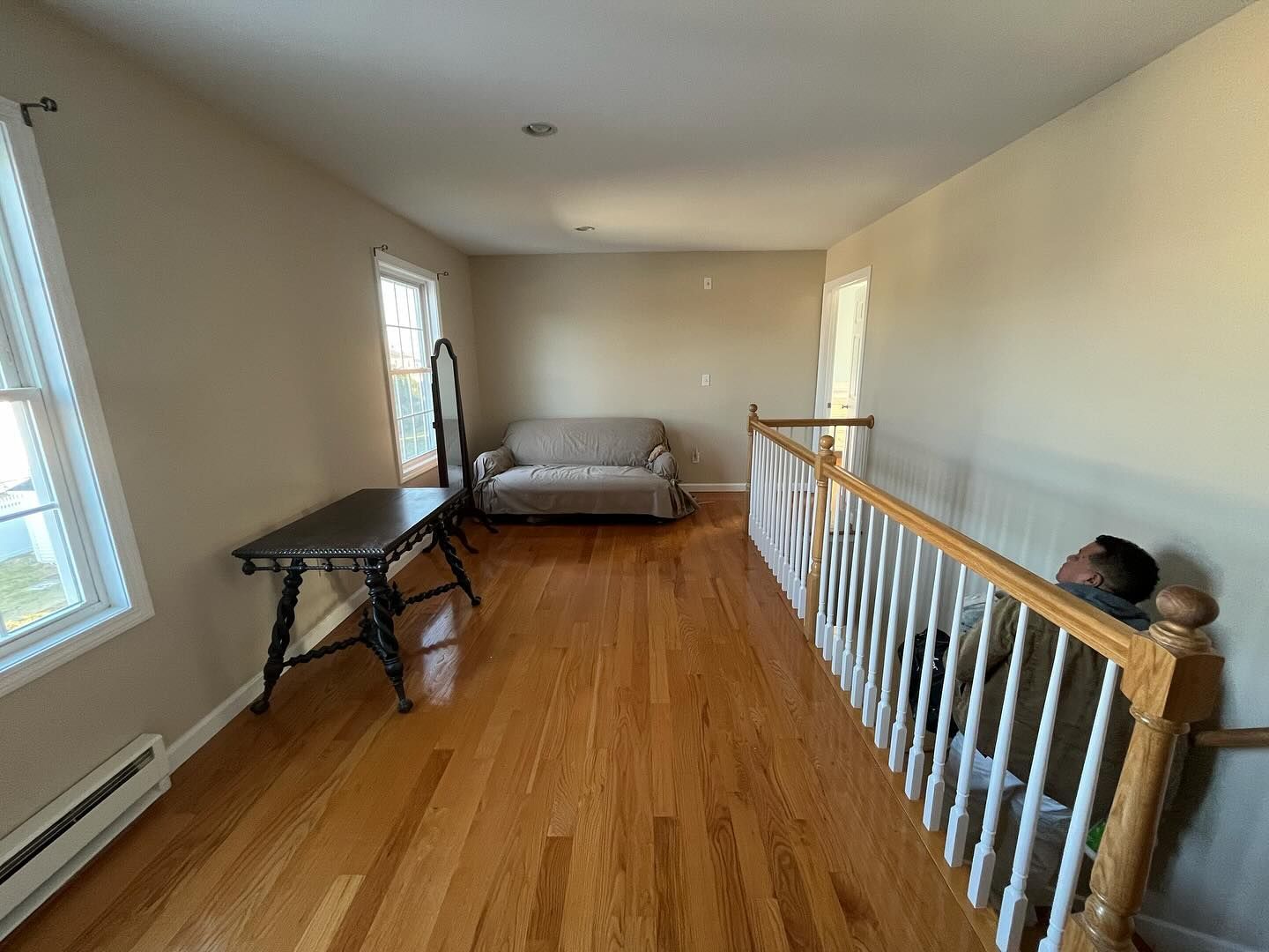 Hallway with hardwood floor, couch, table with mirror, and stair railing. Beige walls, windows.
