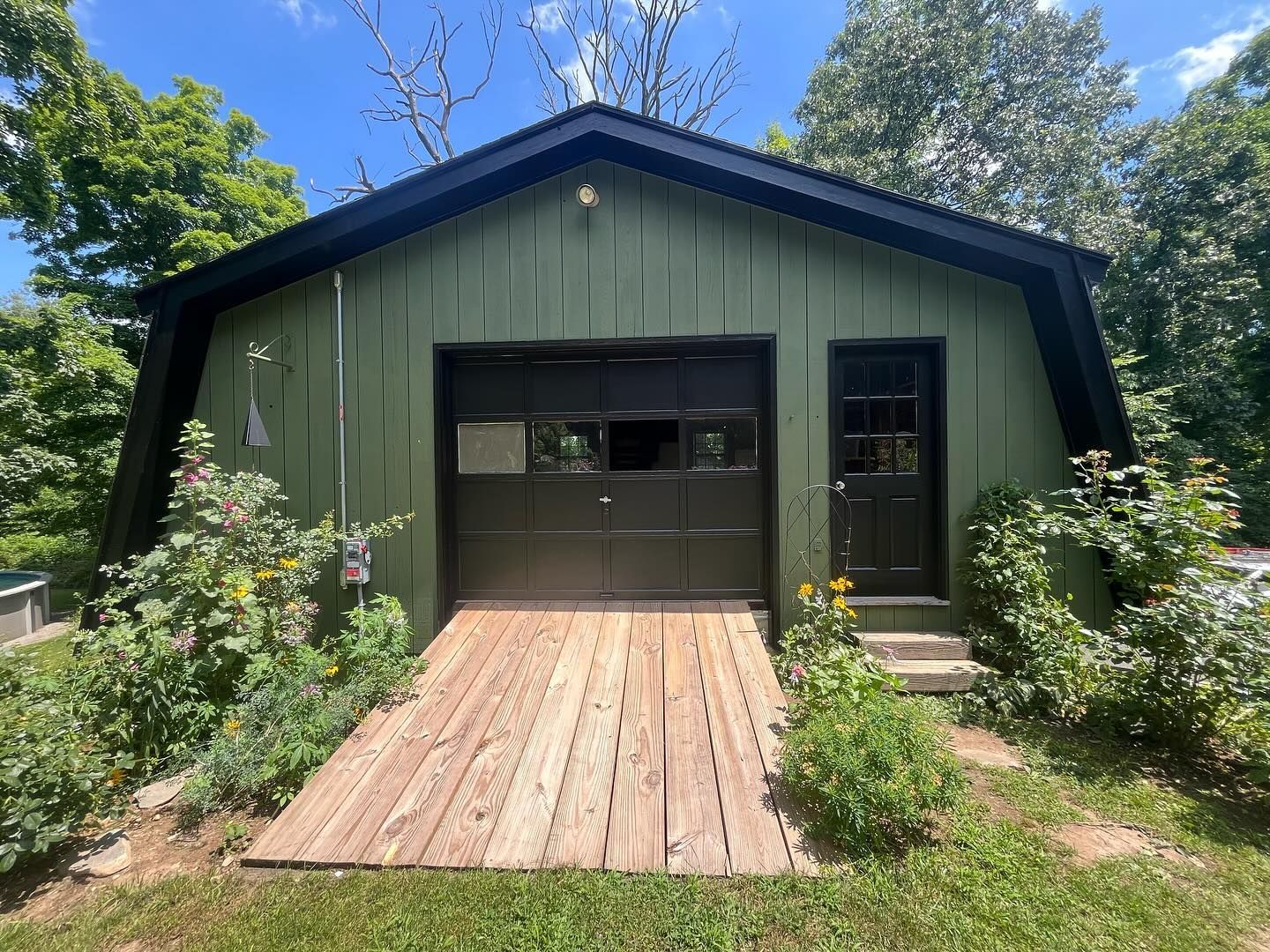 Green garage with black door and trim; wooden ramp and door. Surrounded by greenery and grass.