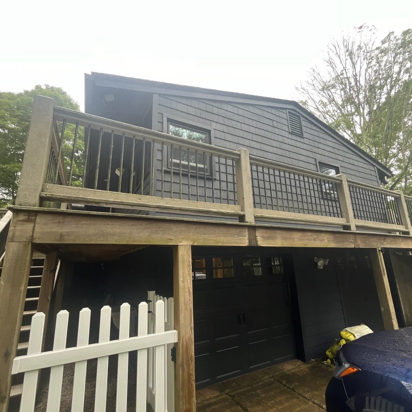 Black house with a deck over a garage. White picket fence in front.