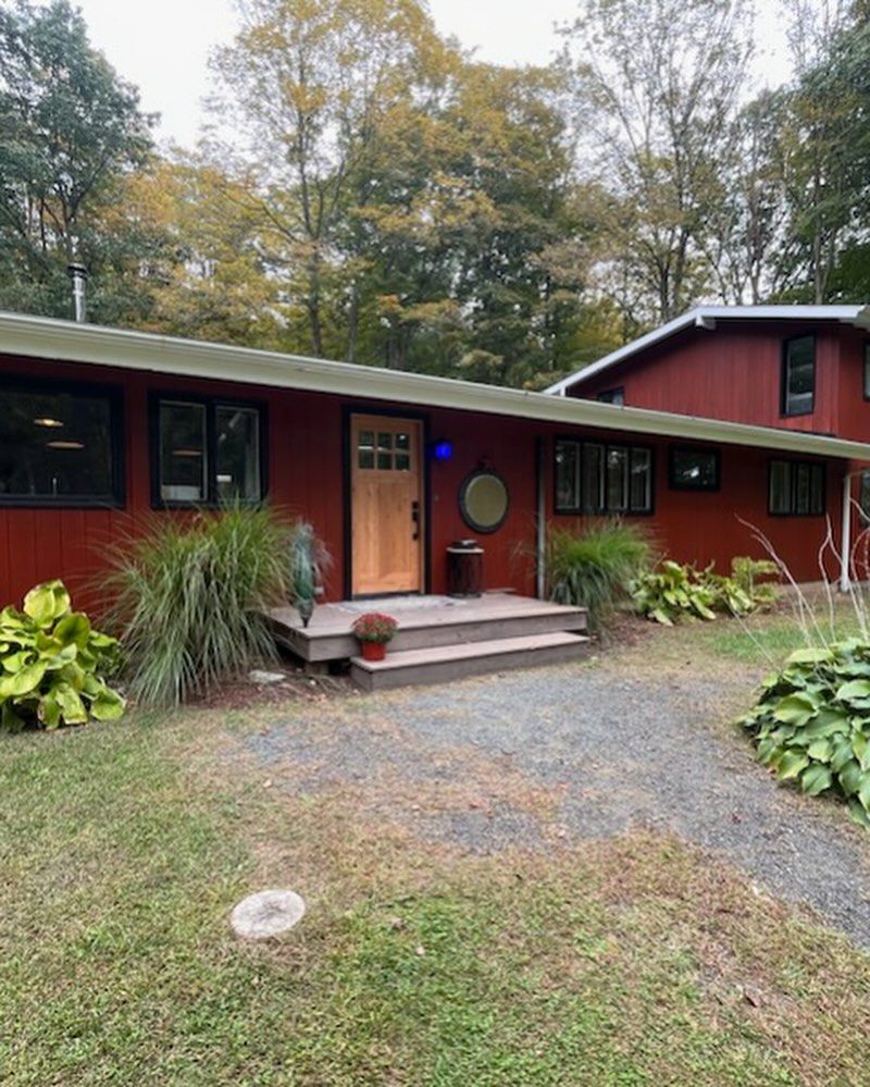 Red house with a wooden door, surrounded by foliage and a gravel path leading to the entrance.
