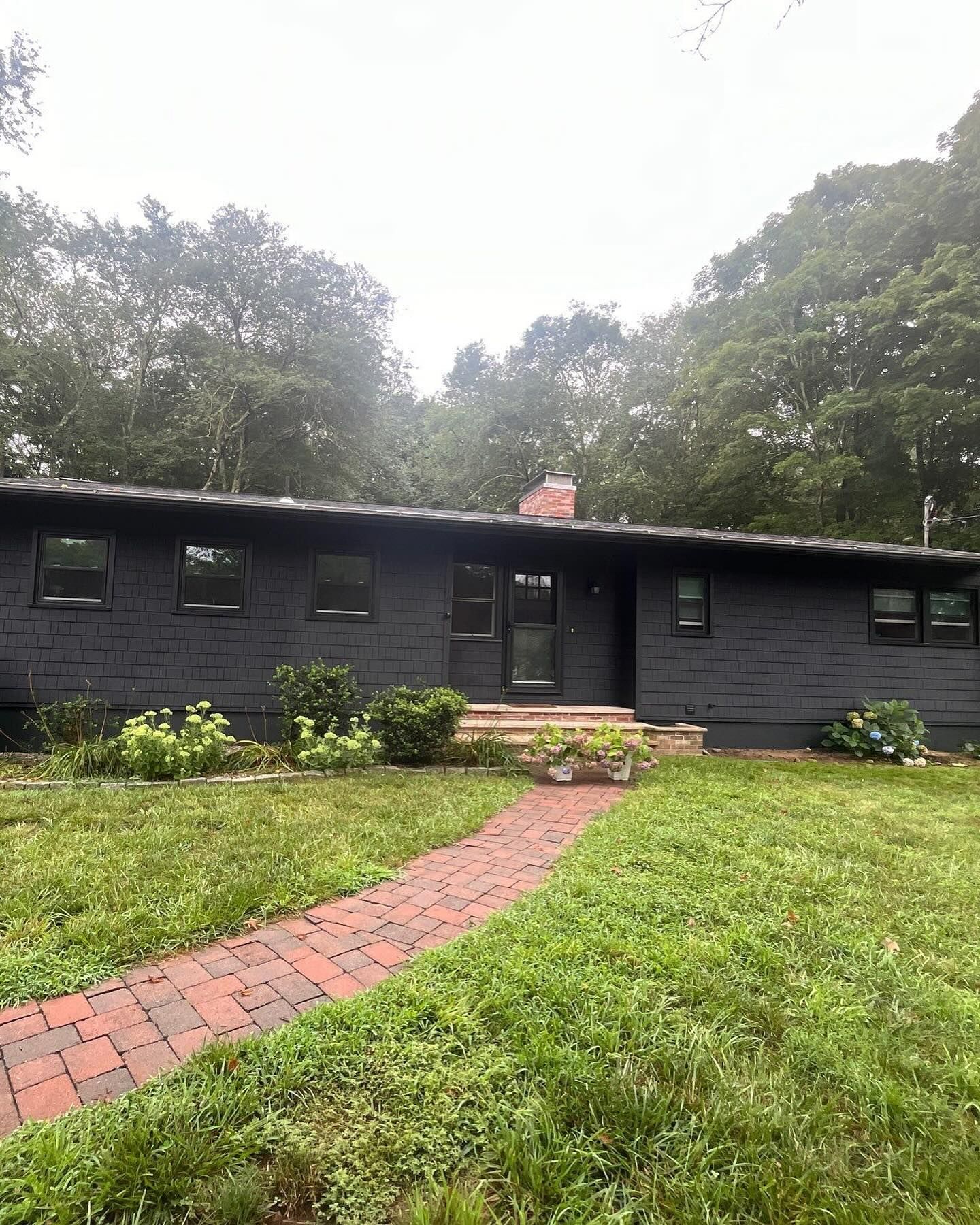 Dark gray house with brick pathway leading to the front door. Lush green lawn and trees in the background.