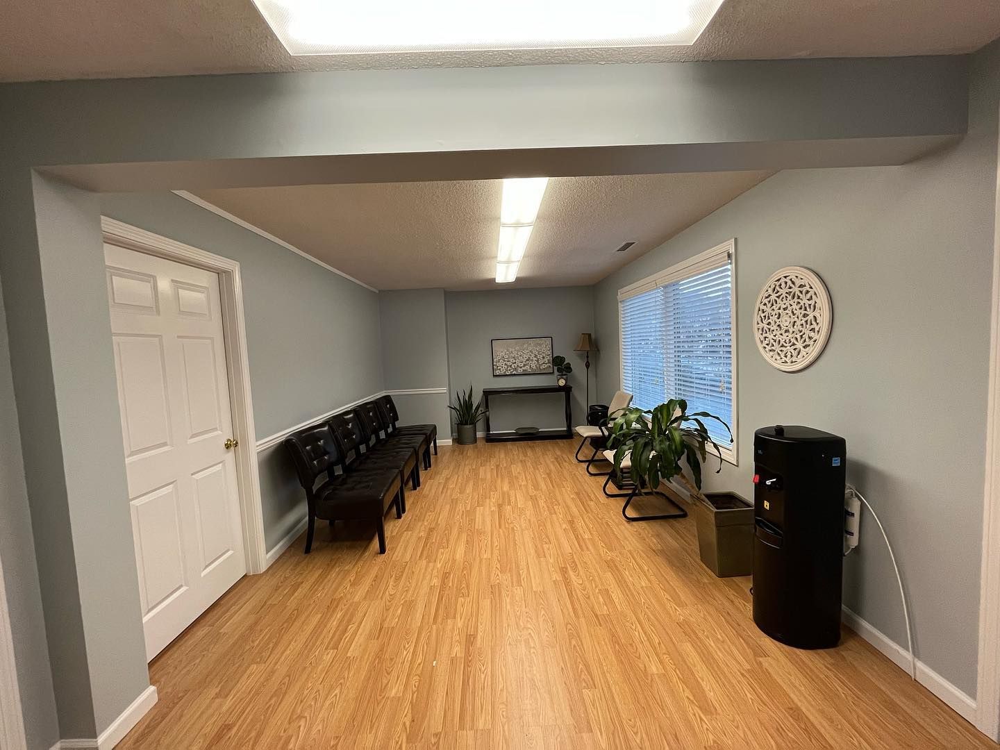 Waiting room with benches, plants, and a water cooler. Light wood-look flooring and light blue walls.
