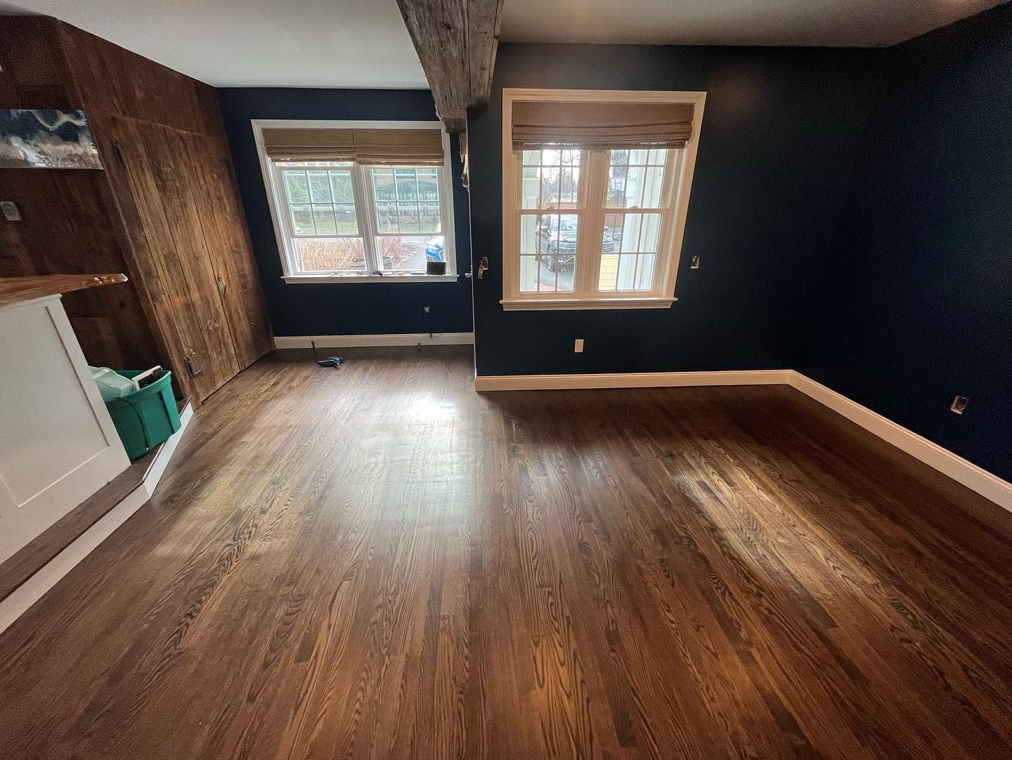 Empty room with dark wood flooring, navy walls, two windows with blinds, and wood paneling.