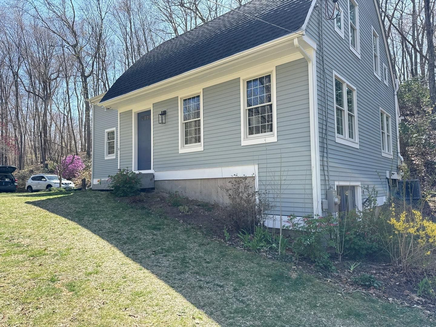 Two-story blue house with white trim, set on a grassy hill; trees in the background.