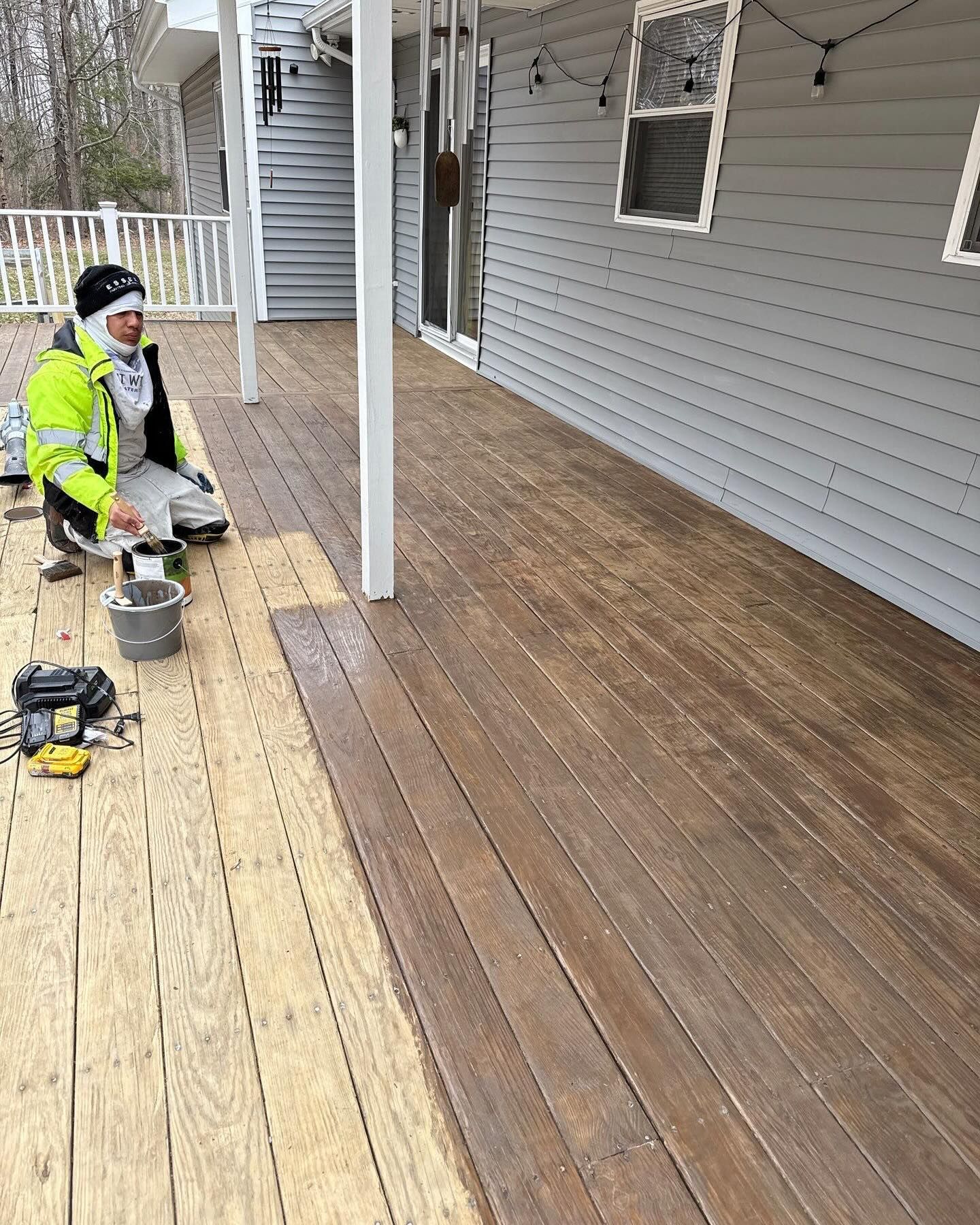 A person on a deck, applying stain. The deck is partially stained, showing a color difference. The siding is gray.