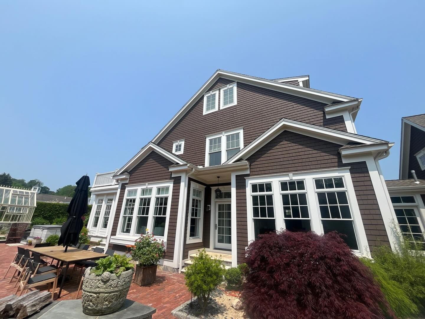 Brown house with white trim, multiple windows, and a brick patio under a clear blue sky.