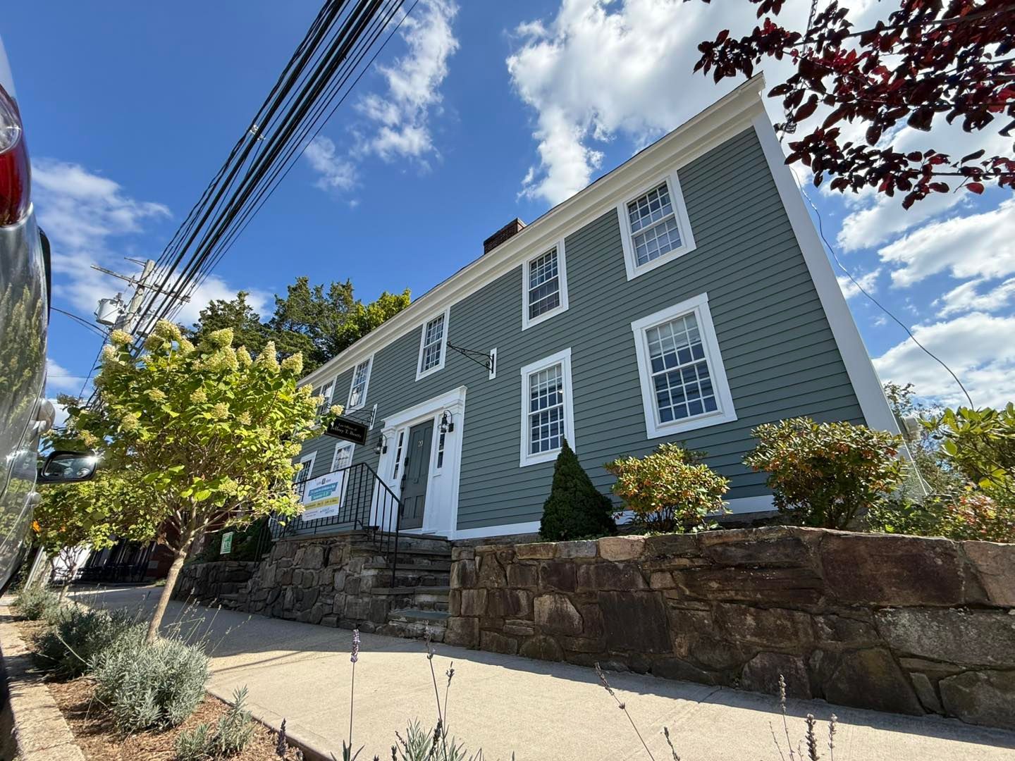 Two-story green clapboard building with white trim, stone wall, and blue sky.