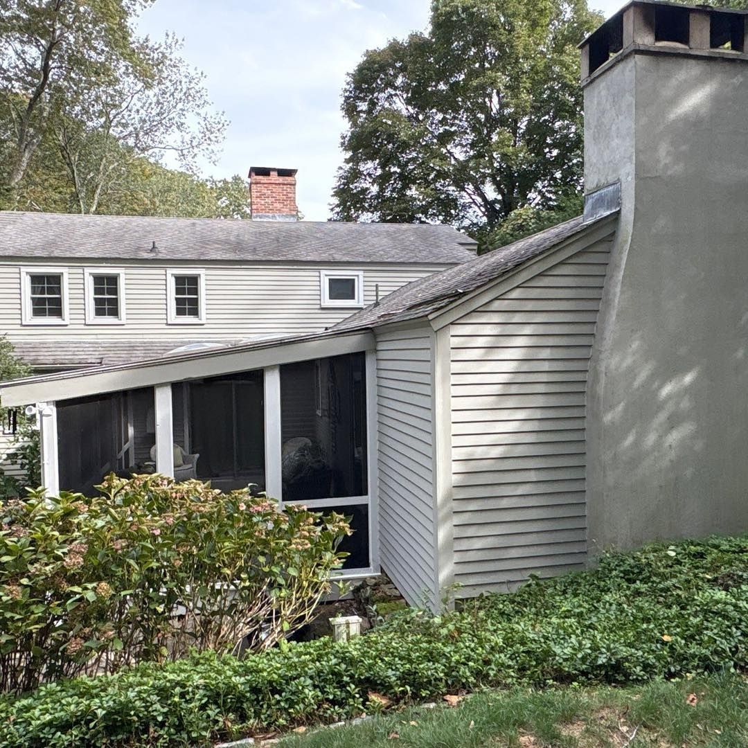 Exterior of a light-colored house with a screened-in porch and chimney, surrounded by green bushes and trees.