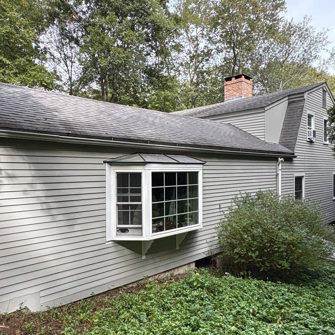 Gray-sided house with a bay window, chimney, and dark roof amidst trees and foliage.