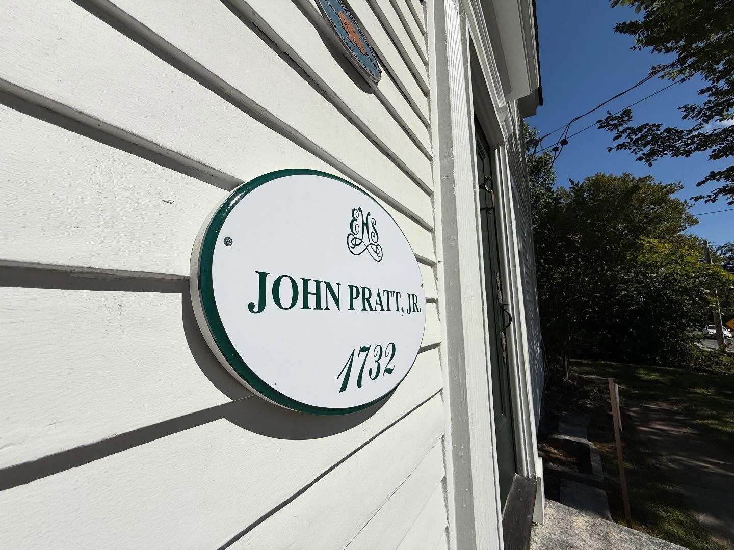 White sign with green trim on a white clapboard wall.
