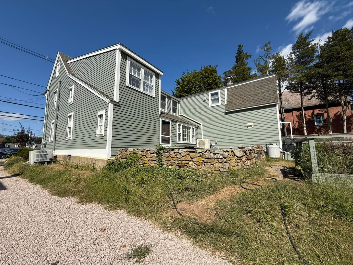 Two-story gray house with white trim, stone wall, and overgrown grassy hill under a blue sky.
