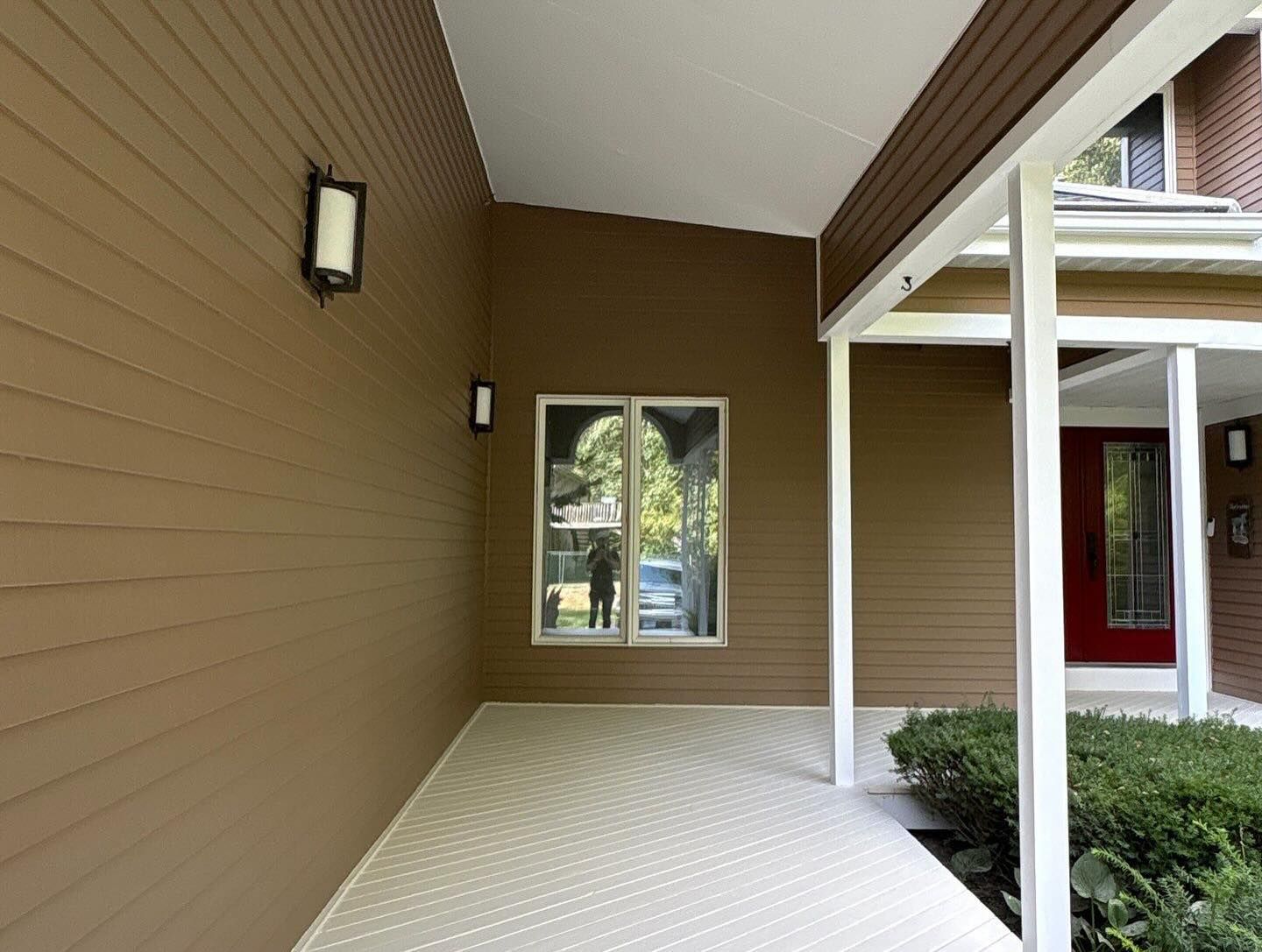 Brown-sided home entryway with a covered porch, window, white columns, and red front door.