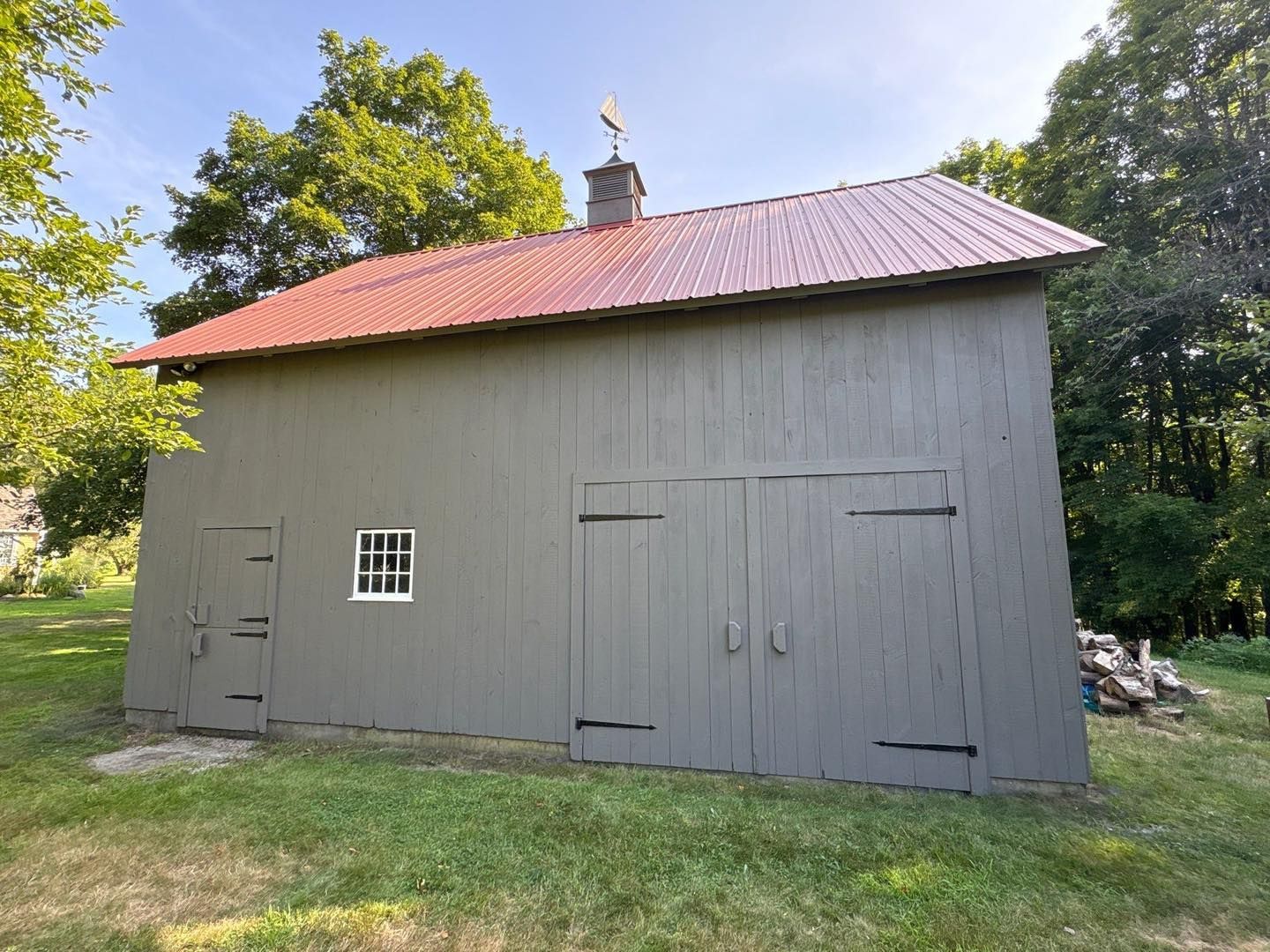 Gray barn with red roof and small window, on a grassy plot with trees in background.