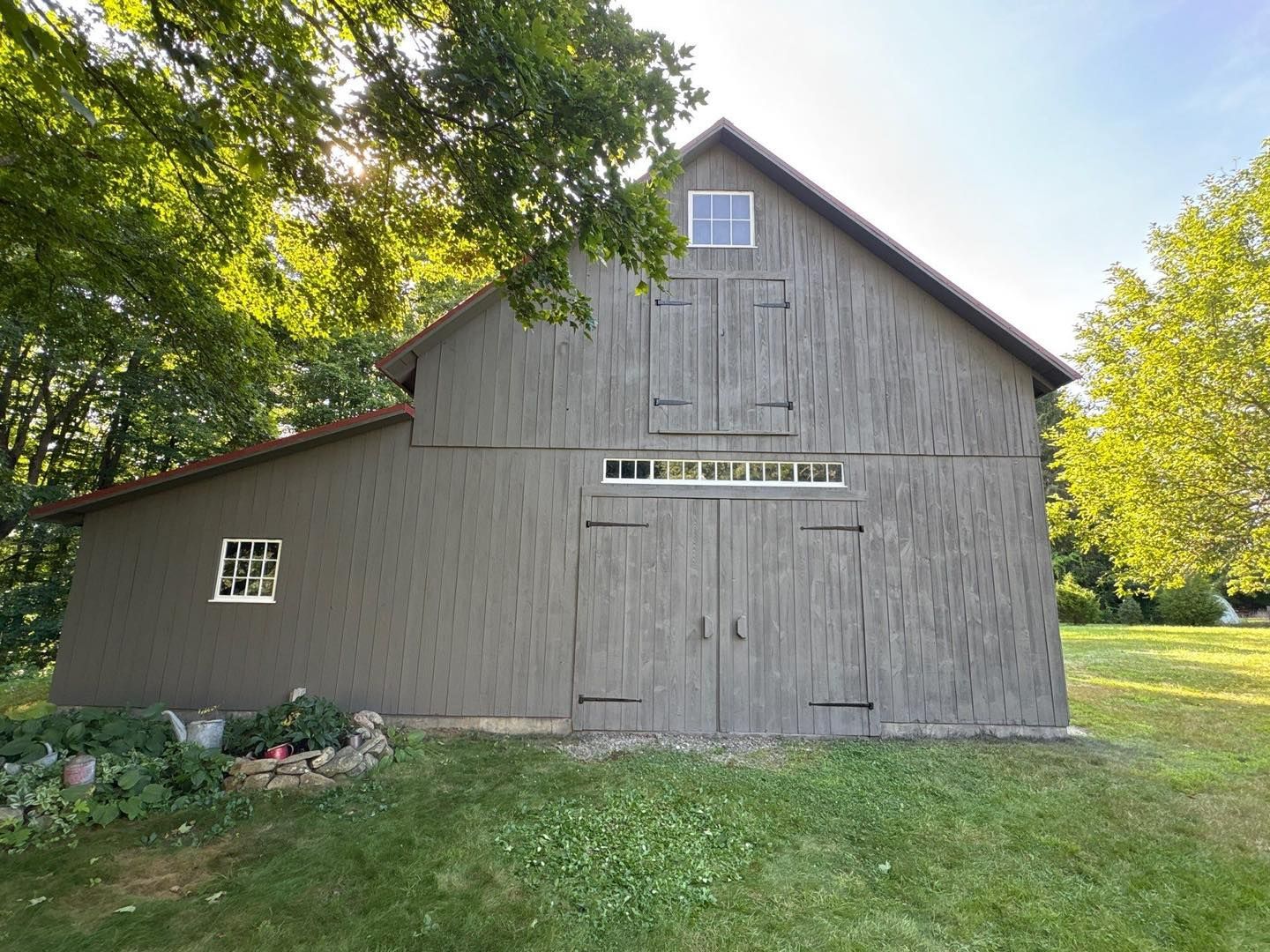 Gray barn with red roof and small windows, set in a grassy yard, partially shaded by trees.
