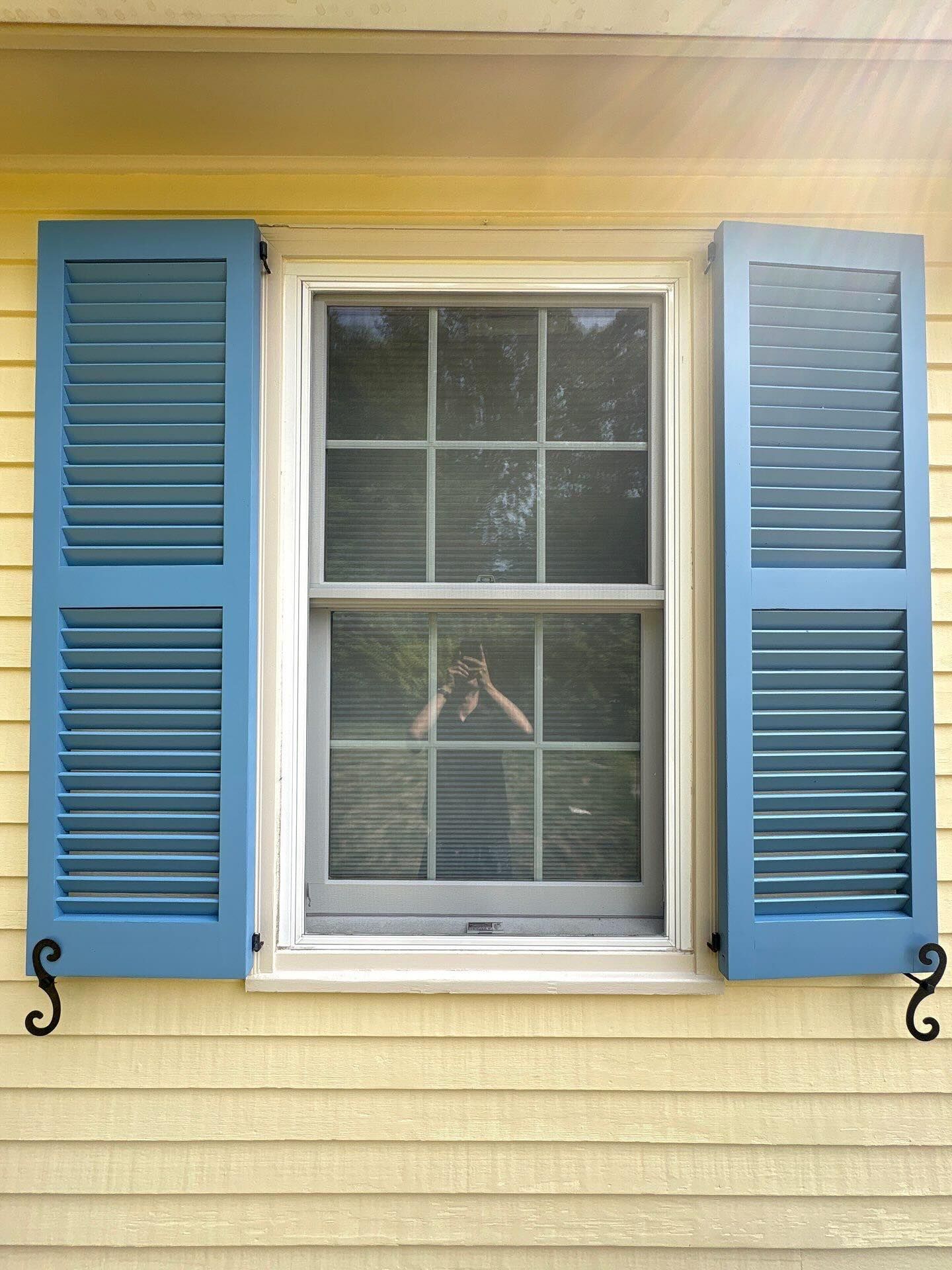 Blue louvered shutters flank a white-framed window on a yellow house. Black hardware.