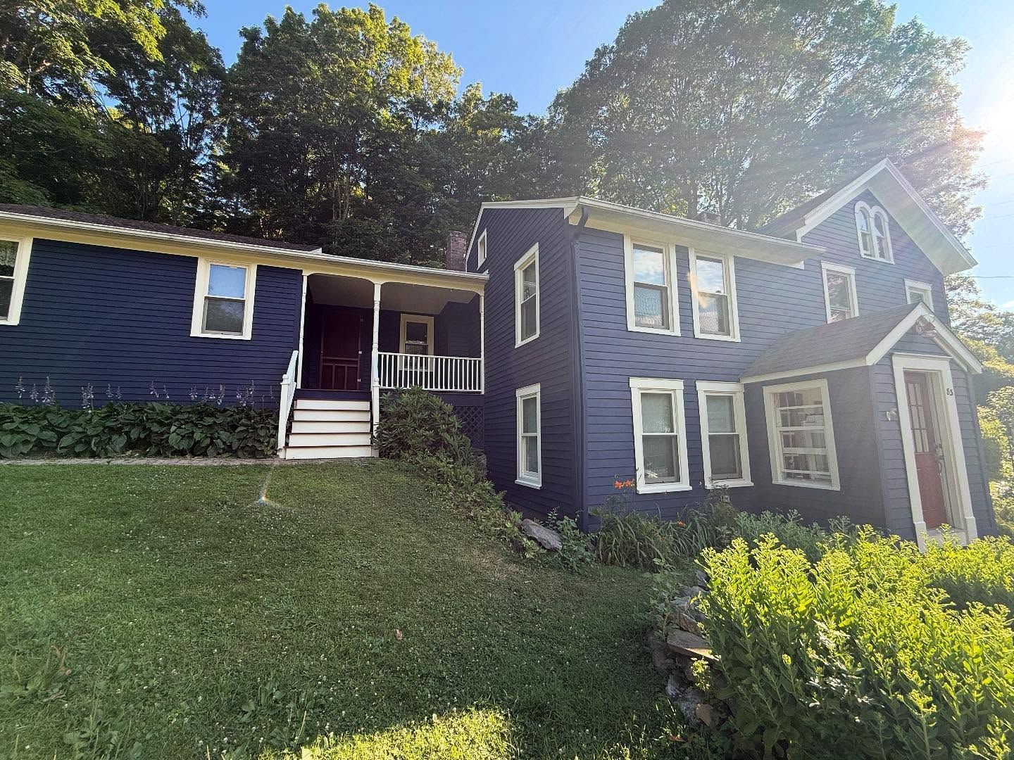 Blue house with porch and white trim nestled in front of a green hillside and trees. Sunny day.