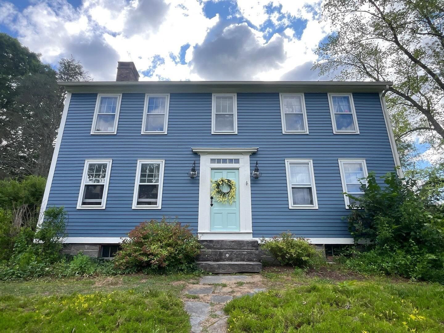 Blue colonial house with white trim, teal door, and wreath, under a cloudy sky.