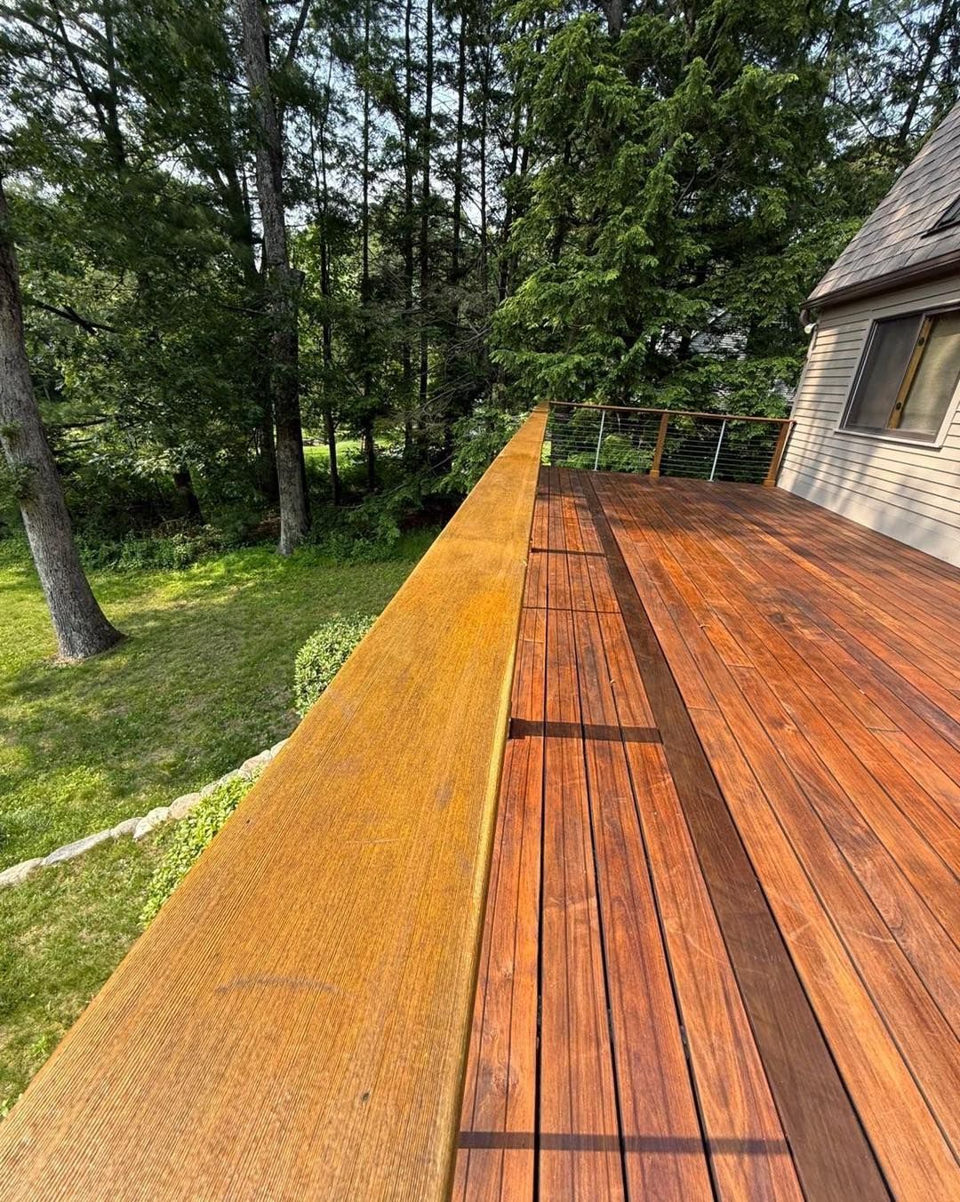 Wooden deck with a brown railing, overlooking a green yard and trees, next to a brick house.