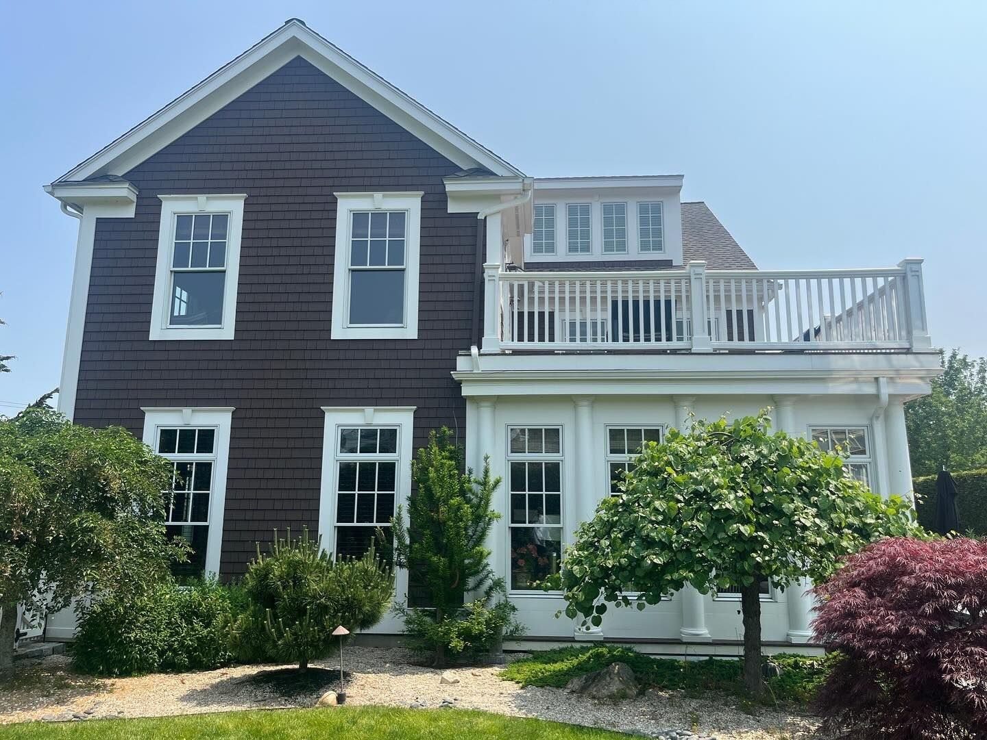 Brown and white two-story house with a deck. The house has a white trim and windows, with shrubs in front.