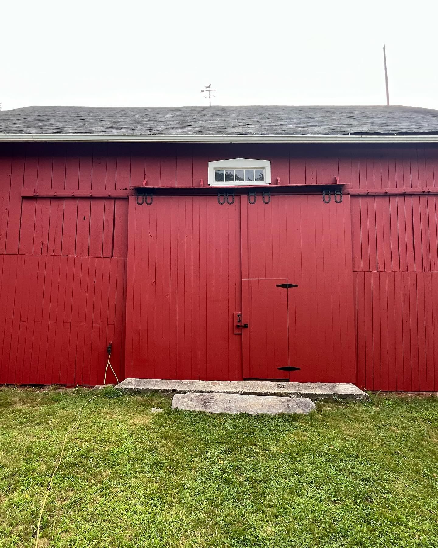 Red barn with large sliding doors, small door within, white window, and a gray concrete step.