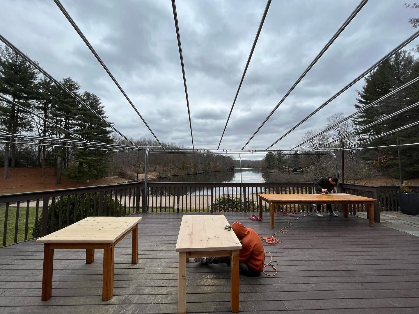 Wooden tables on a deck with people working, overlooking a river. Overhead cables run across a cloudy sky.