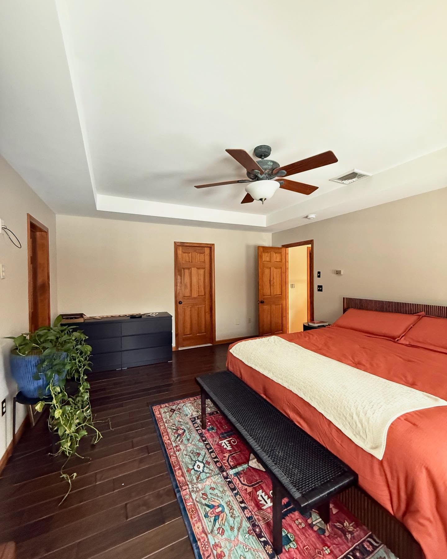 Bedroom with dark wood floors, orange bedding, black bench, and a red and blue rug.