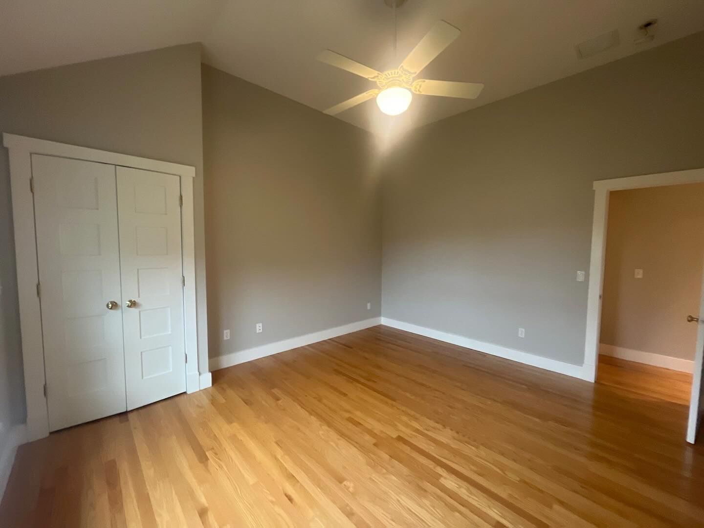 Empty room with hardwood floors, white closet, and doorway. Beige walls, ceiling fan, natural light.