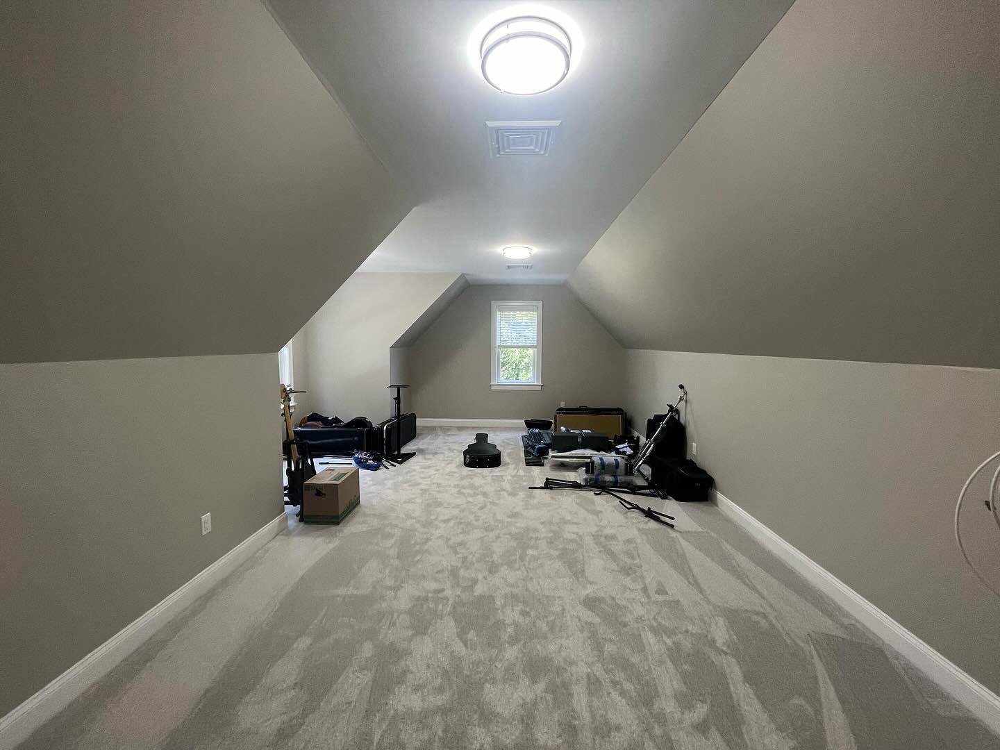 Spacious attic room with gray carpet and walls. Ceiling lights, window, and clutter in the center.