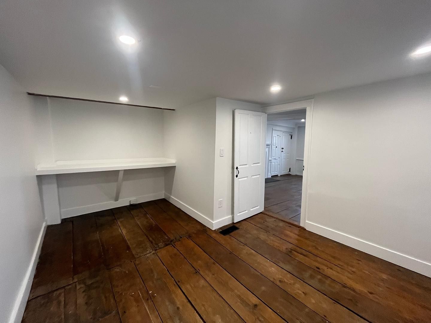 Room with wood floor, white walls, built-in desk, and doorway to another room.