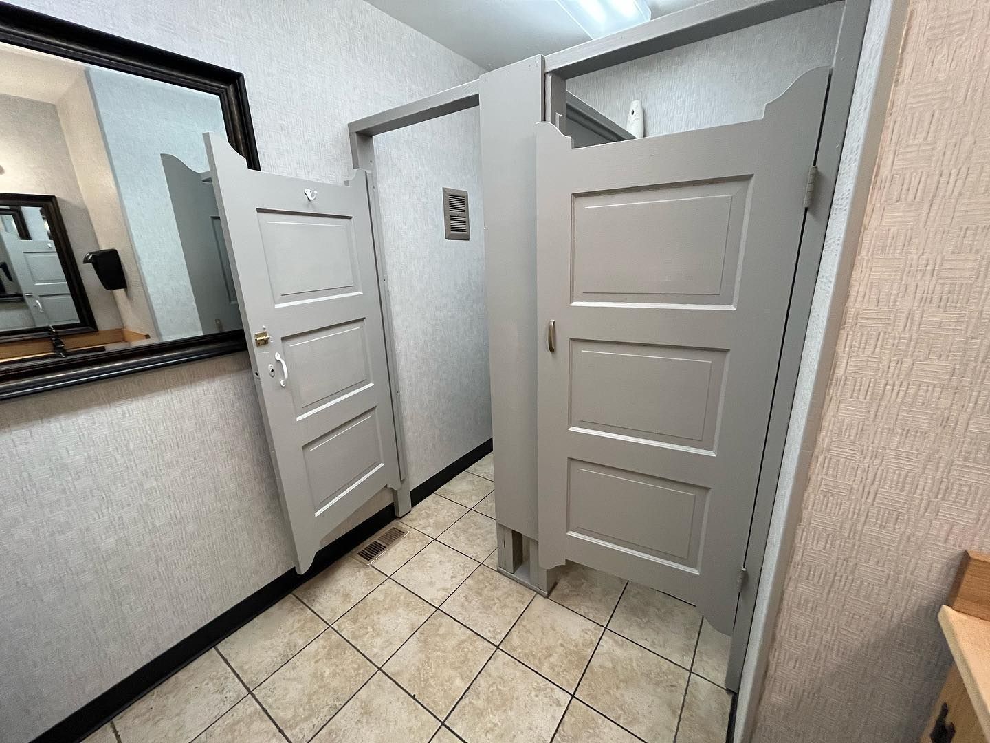 Bathroom stall doors, gray, ajar, over tiled floor. Mirror and textured walls visible.