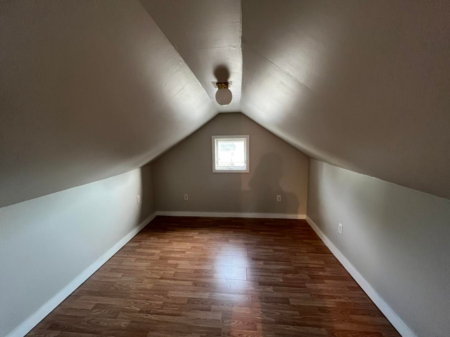Empty attic room with wood-look flooring, sloped walls, small window, and light fixture.