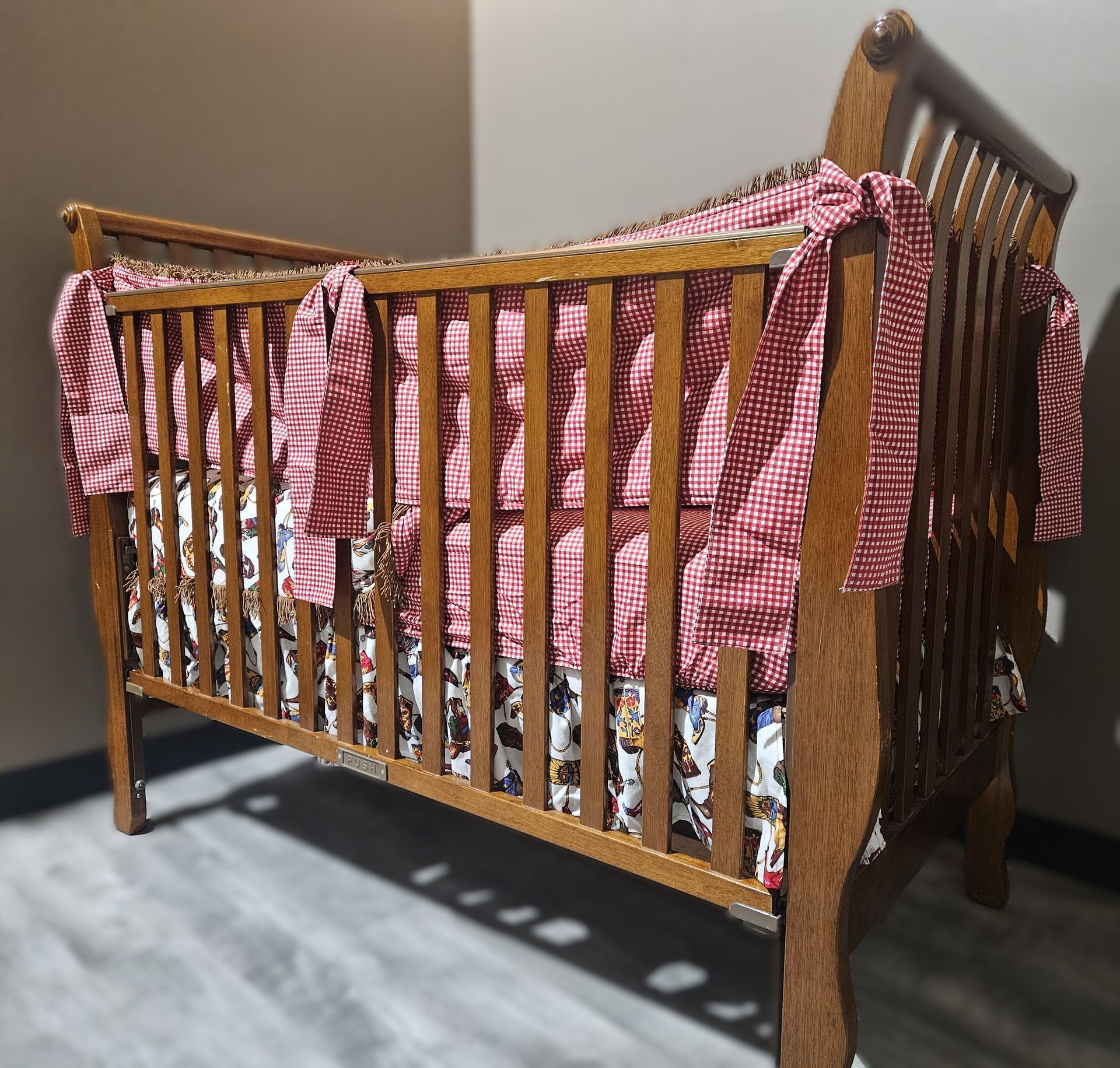 A wooden crib with a patterned skirt and a red and white checkered bumper tied to the slats.