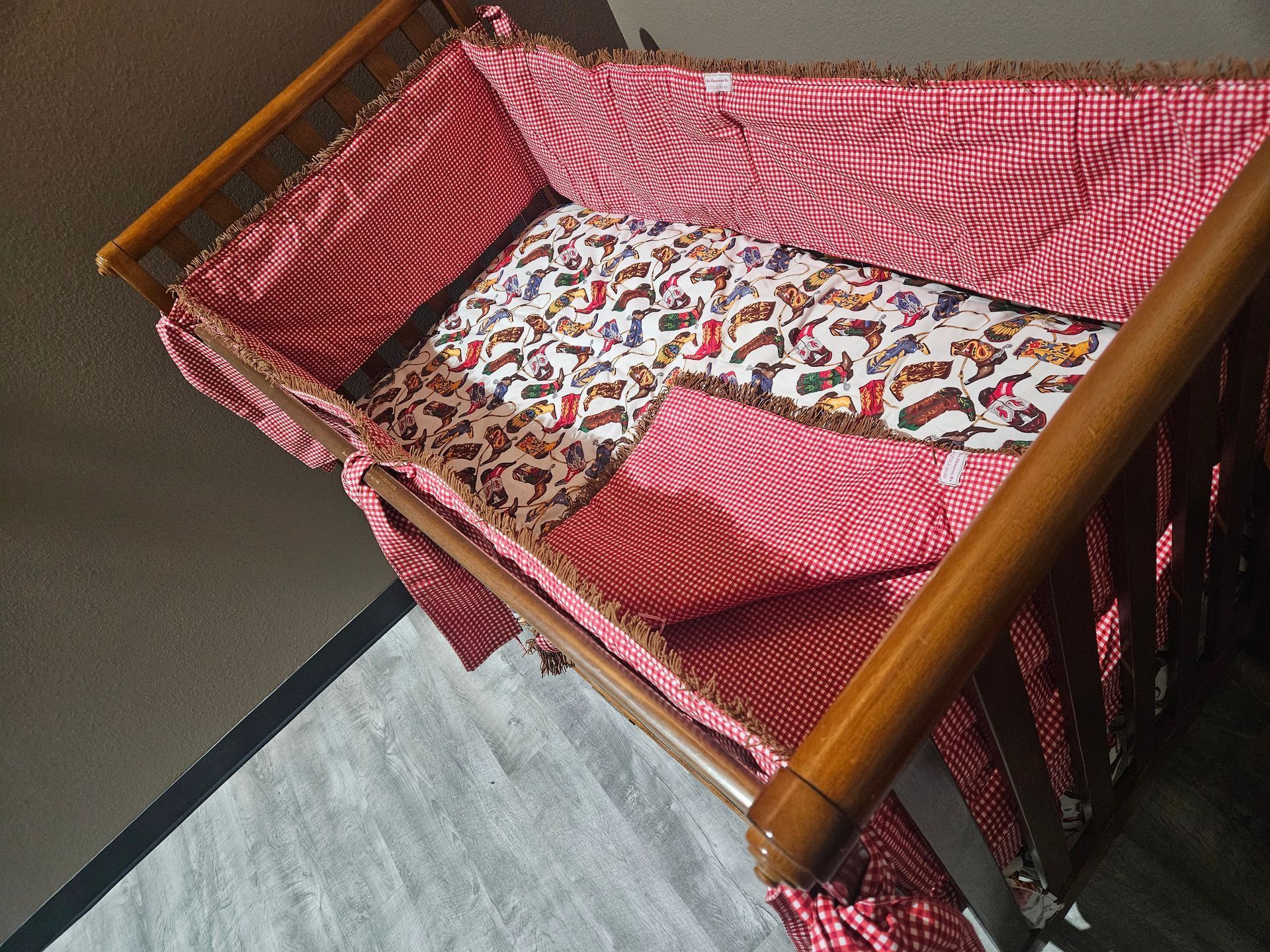 A wooden baby crib with a red-and-white checkered bumper pad and a patterned crib sheet featuring various animals.