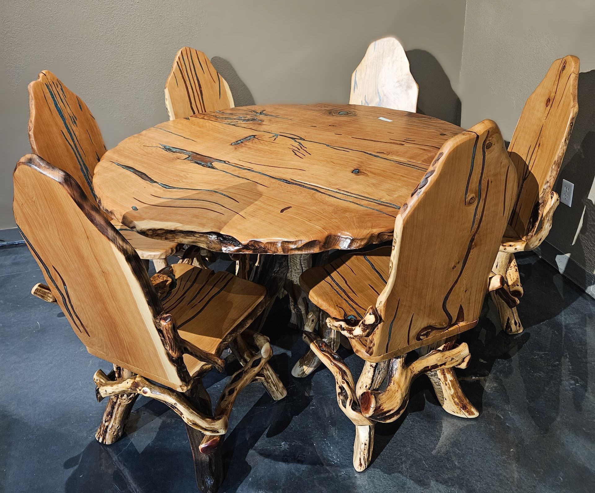 A rustic, circular wooden dining table with live-edge details surrounded by five matching chairs on a dark, glossy floor.