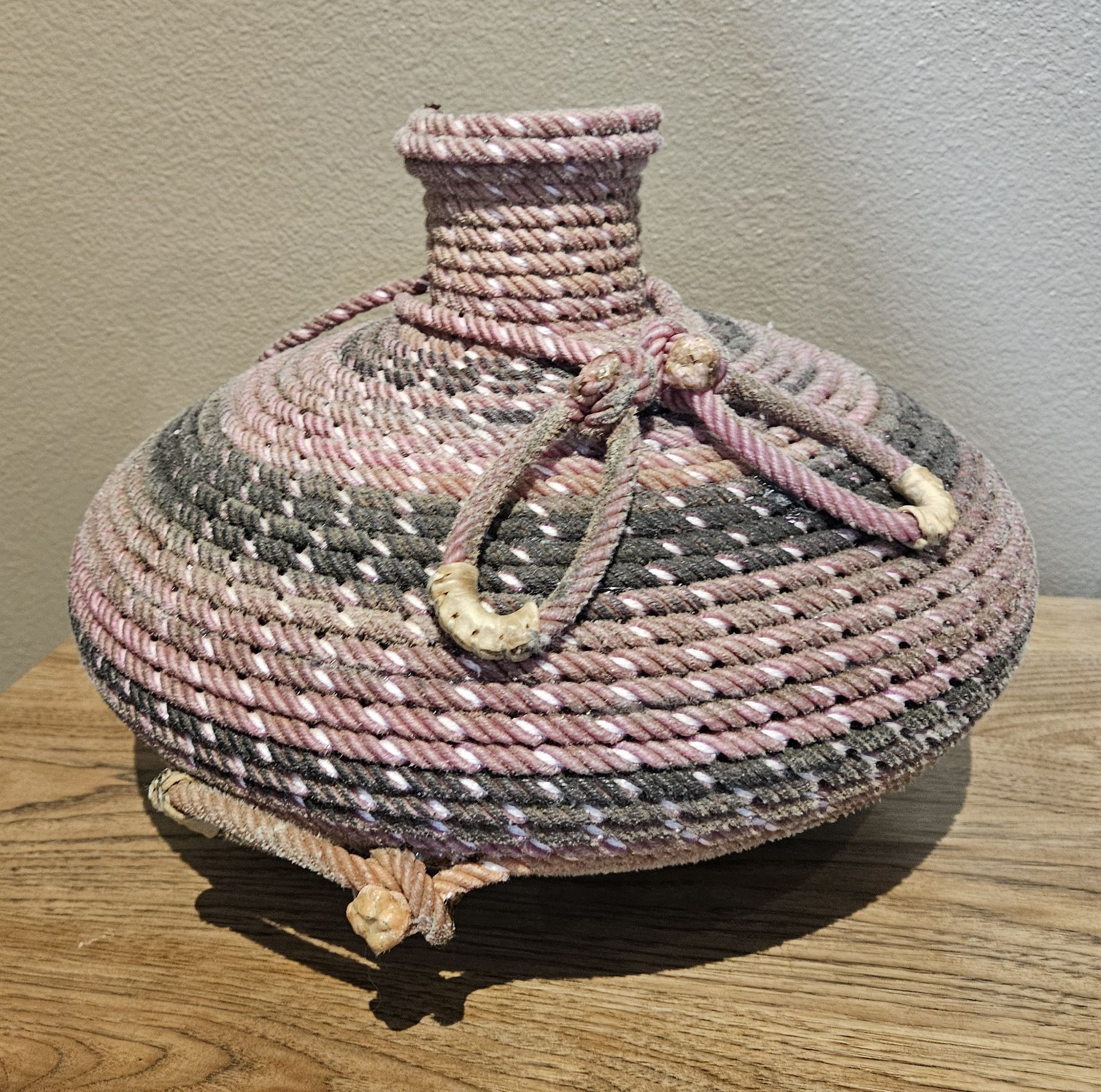 A rounded vase made of coiled pink, grey, and white rope with decorative looped handles, sitting on a wooden surface.