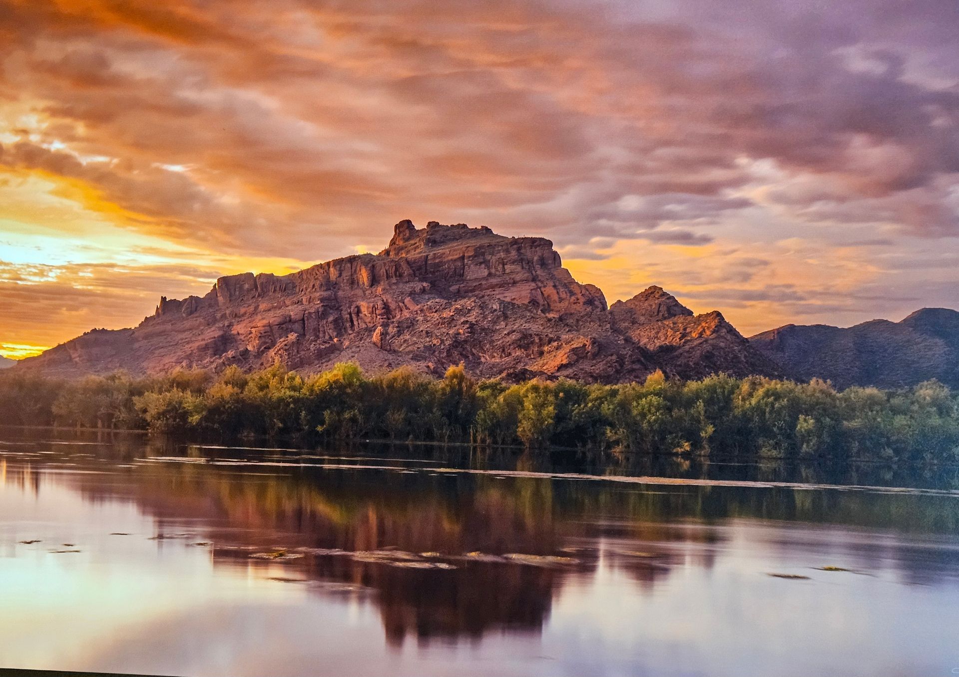 A rocky mountain range reflecting in calm water at sunset, with golden and purple clouds filling the sky above.