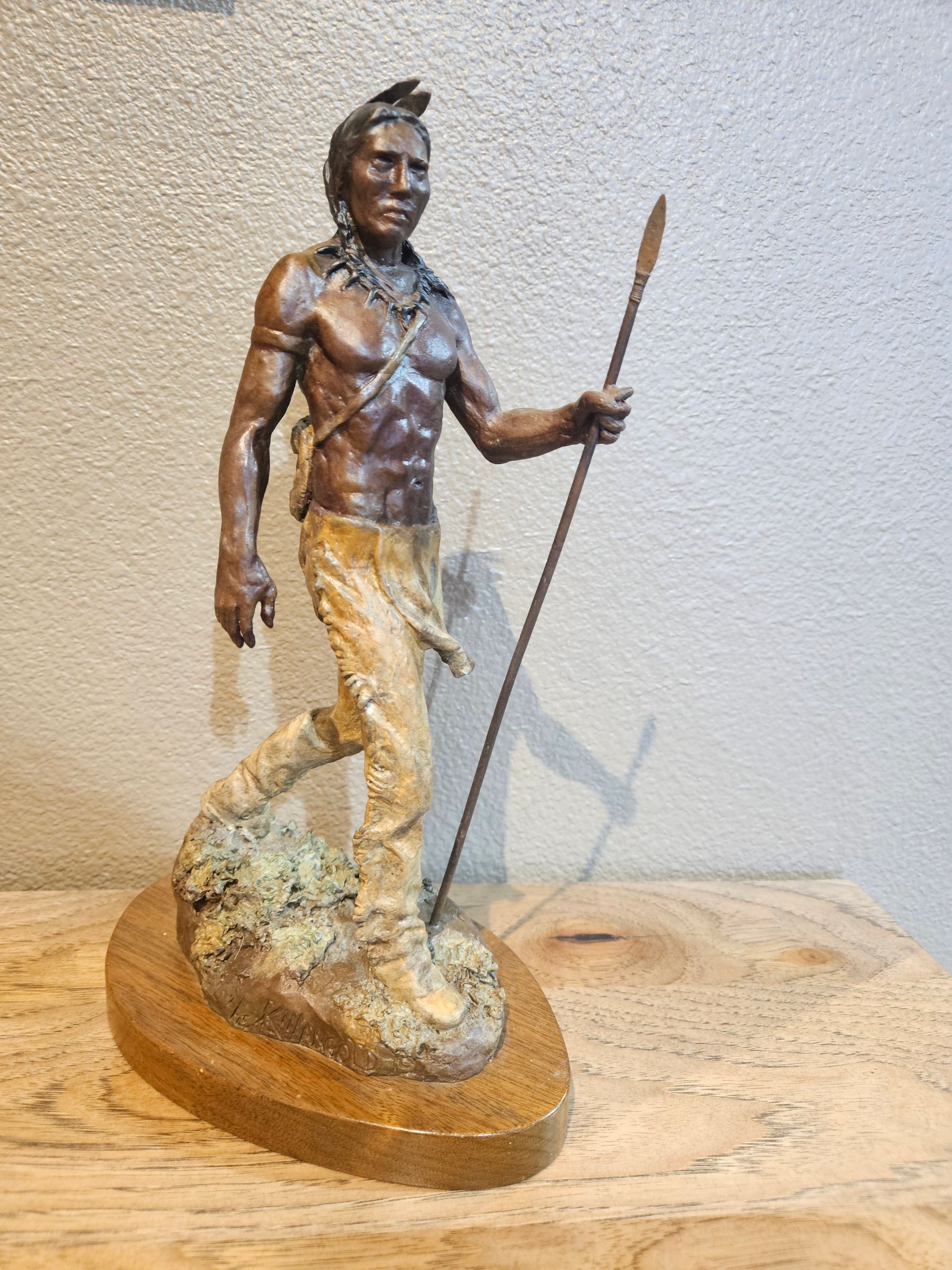 A bronze sculpture of a figure wearing fringed pants and holding a spear, standing on a rocky base.