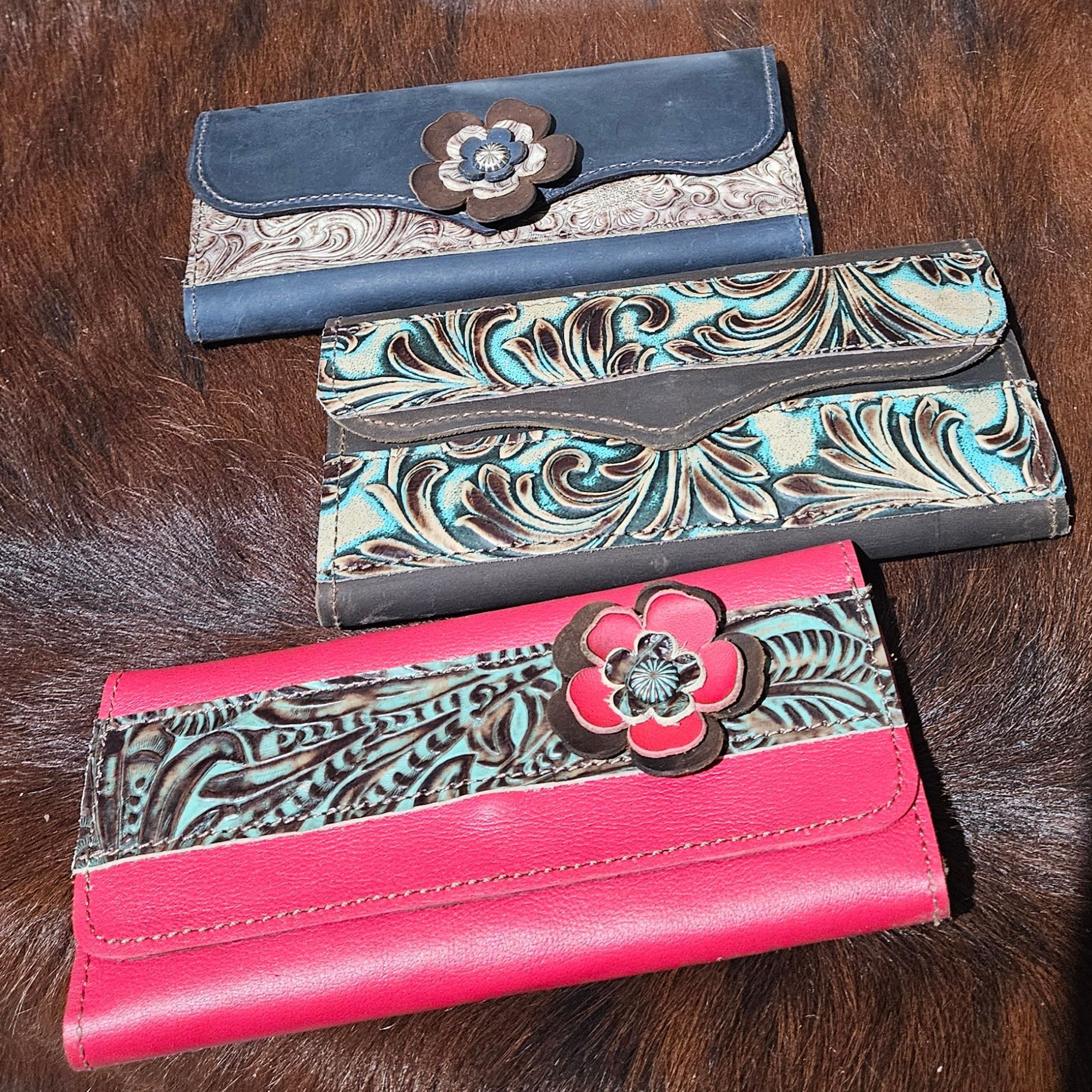 Three tri-fold wallets in pink, turquoise-patterned leather, and blue, each featuring a decorative leather flower detail.