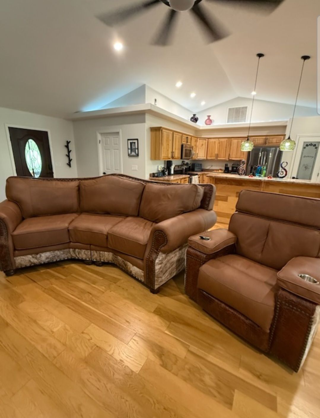 Living room with brown sectional and recliner, hardwood floor, open kitchen, and ceiling fan lights.