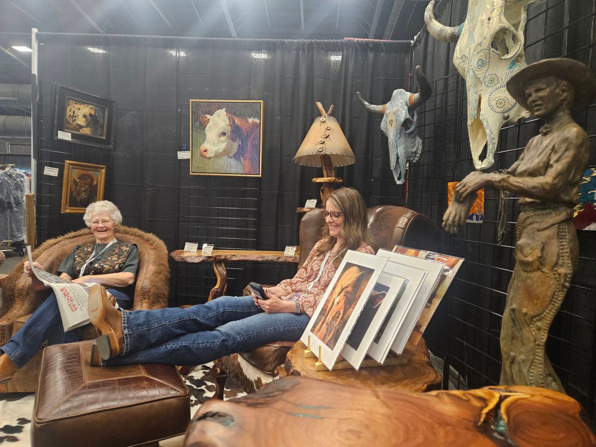 Two people in a western-themed art booth. One reads, the other looks at a tablet. Artwork and sculptures are visible.