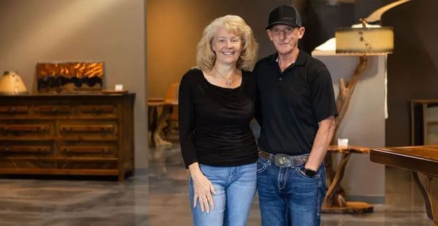 Couple standing inside a room with furniture, woman in black top and jeans, man in black shirt and hat.