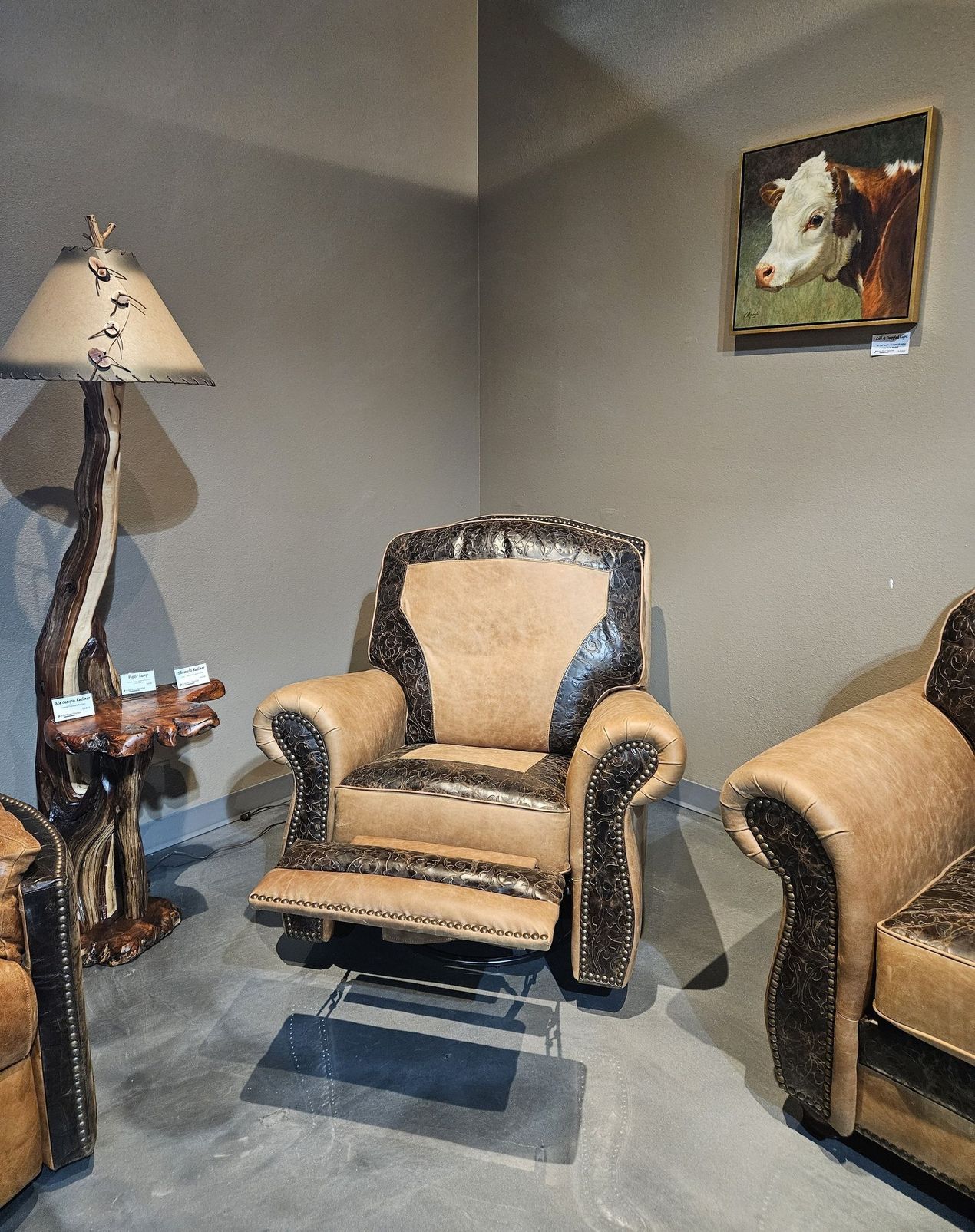 A room with a rustic recliner, a cow-print accent chair, a matching lamp with a base, and a small cow portrait on the wall.