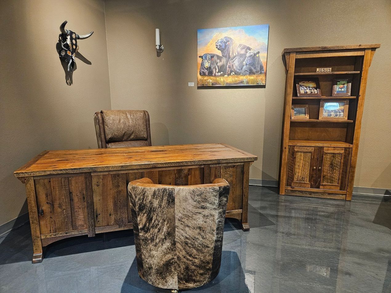 A rustic office features a wooden desk and chair, a matching bookshelf, a buffalo painting, and a steer skull wall decor.