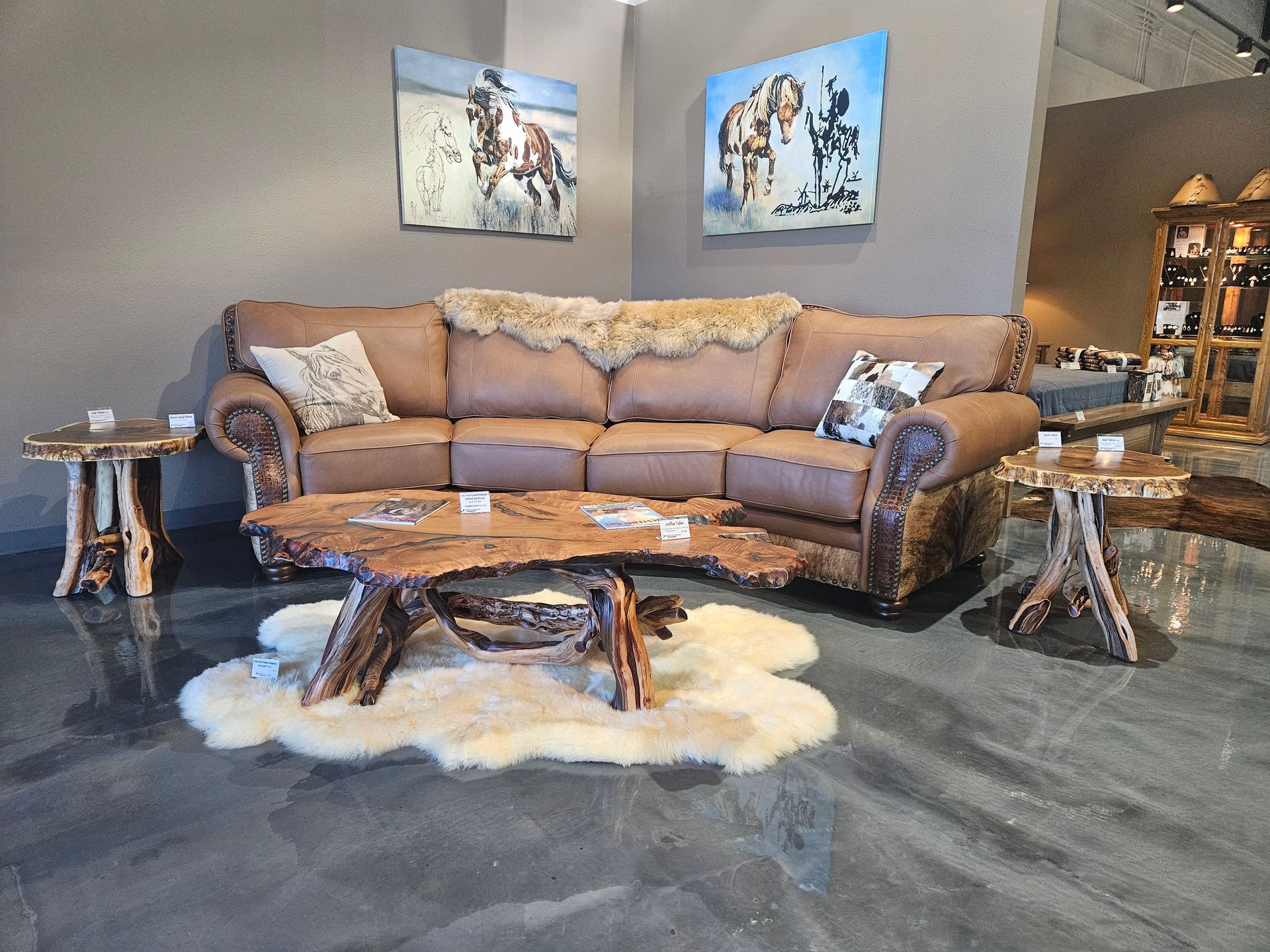A tan leather sofa with rustic log tables and animal hide rugs on a polished concrete floor in a furniture showroom.