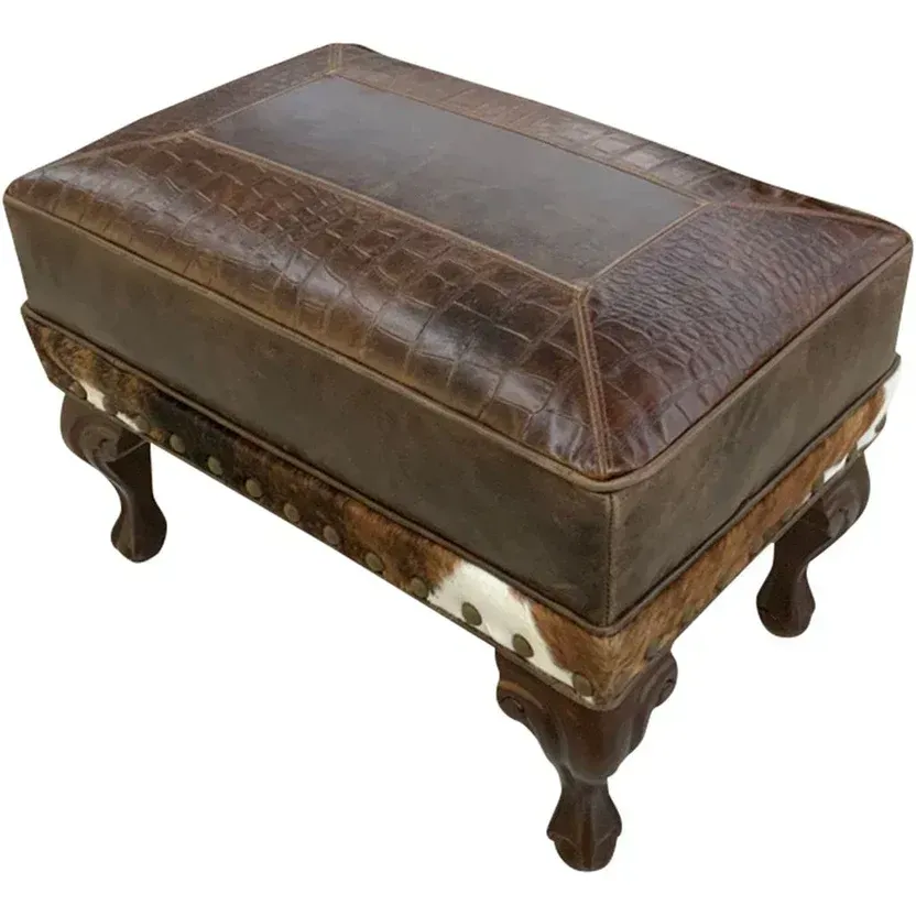 A brown leather footstool with crocodile-embossed top, cowhide sides, and dark ornate wooden legs.