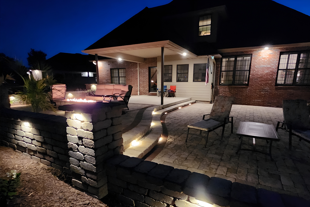 Lit garden path beside a brick house, with flowers, shrubs, and a tree at dusk